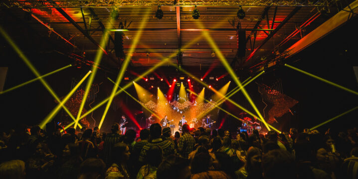 Fra konsert. Mange publikummere med ryggen til i forgrunnen. Gule laserlys på scenen og et band som spiller.