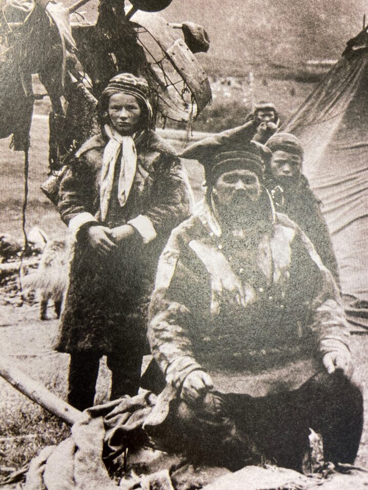 Gammelt bilde av samer på sommerboplassen på Eidebakken. Mann med kofte sitter i forgrunnen. To koftekledde står bak han.