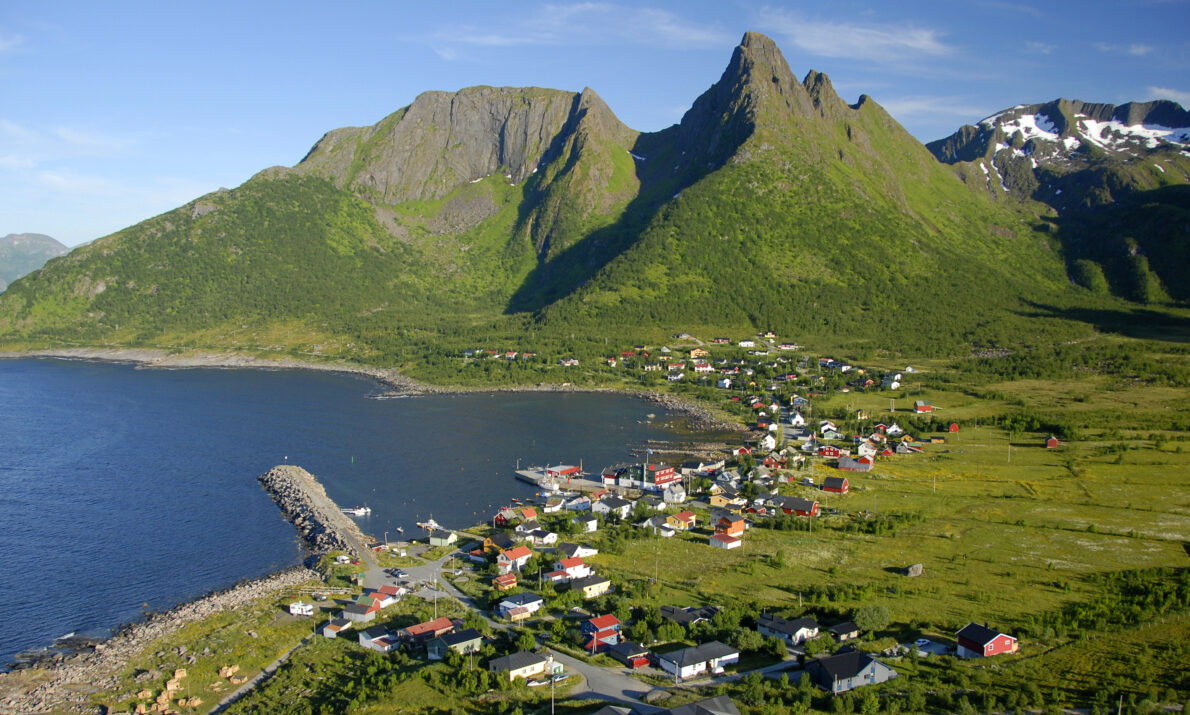 Fiskevær innerst i en liten fjord sett ovenfra. Spisse grønne fjell i bakgrunnen og knall blå himmel.