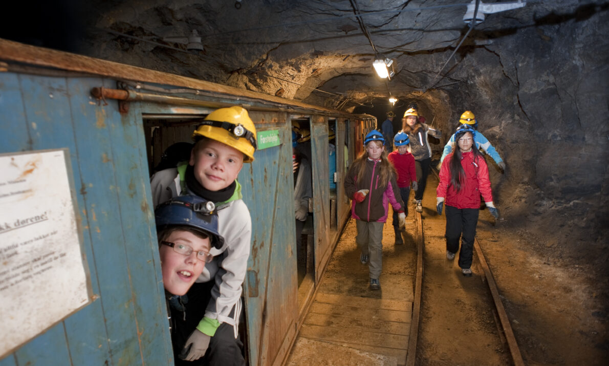 Flere barn inne i en gruve. Non går mot kamera og et par stikker ut fra et lavt, blåmalt bygg inne i gruva. Alle har på hjelm.