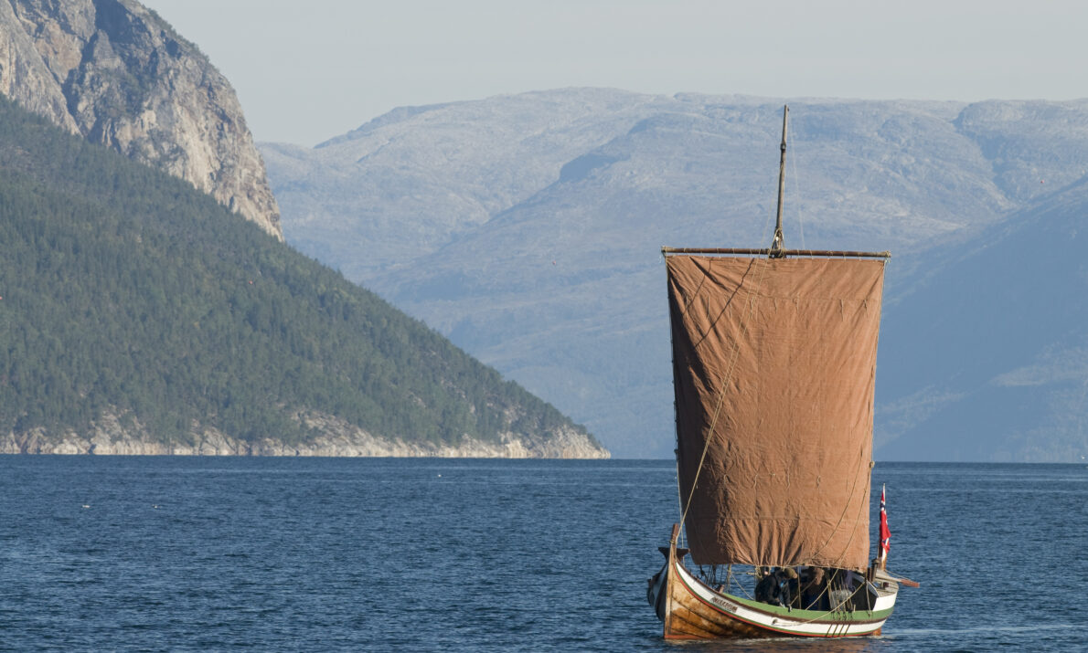 "En tradisjonell trebåt med brun seil seiler på stille vann, med majestetiske fjell og skogkledde åser i bakgrunnen under klar himmel."