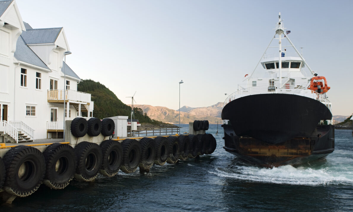 Stor bilferge kommer mot på vannet på vei mot kai. Kai dekket av bildekk. Stor hvit brygge på land. Fjell i bakgrunnen og blå himmel.