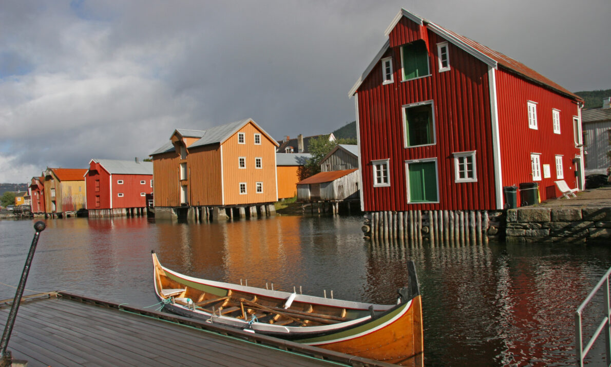 En nordlandsbåt ligger bed brygge på fjorden i forgrunnen. Bak dere en rekke med gamle brygger i rødt og oker.