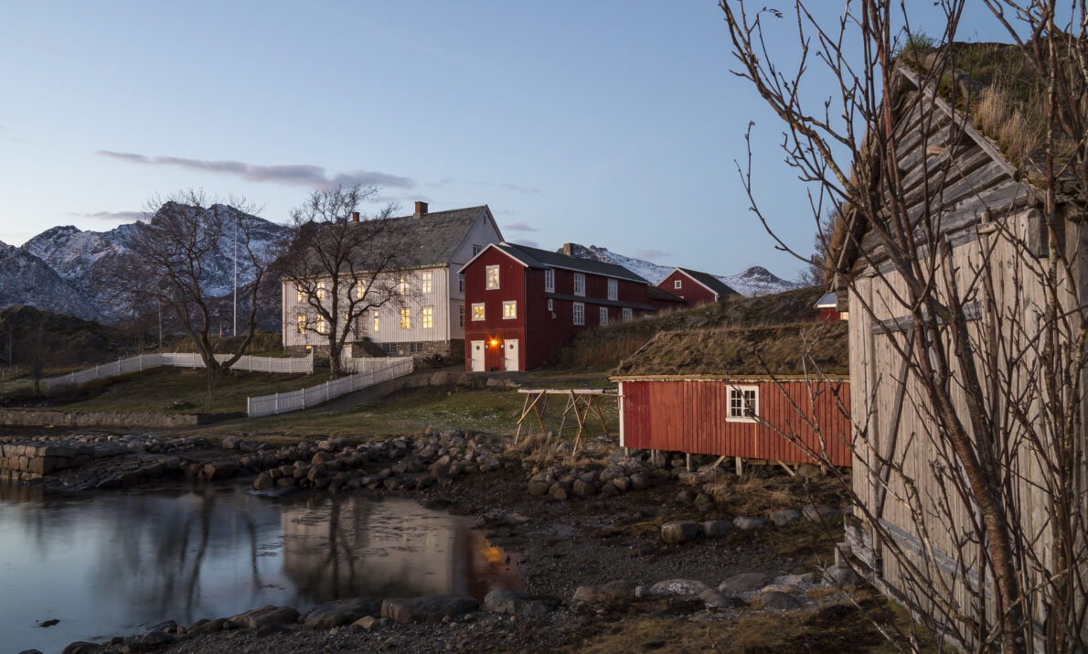 Bildet viser et rolig landskap ved vannkanten i skumringen. Flere hus i røde og lyse farger ligger spredt langs bredden, med fjell som reiser seg i bakgrunnen. Vannet i forgrunnen speiler husene og den dempede himmelen, og trær uten blader står rundt bebyggelsen.