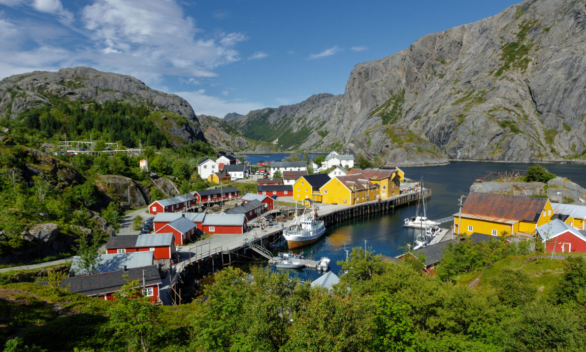 Bildet viser et lite fiskevær omgitt av bratte fjell og grønn vegetasjon. Fargerike bygninger, blant annet flere gule og røde hus, ligger tett rundt en havn med båter og brygger. Vannet er rolig, og himmelen er blå med spredte skyer.