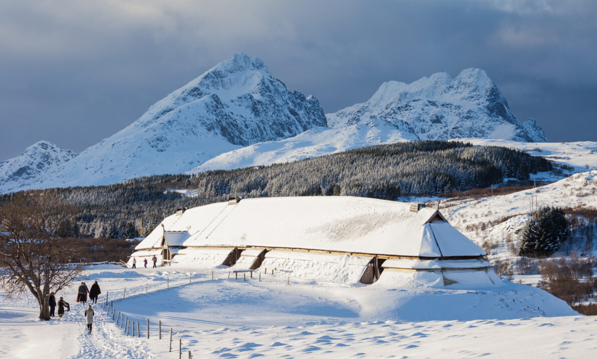 Bildet viser et snødekt vinterlandskap med et snødekt langhus omgitt av gjerder i forgrunnen. To personer går langs en brøytet vei i snøen. I bakgrunnen ruver høye, snødekte fjell under en overskyet himmel.
