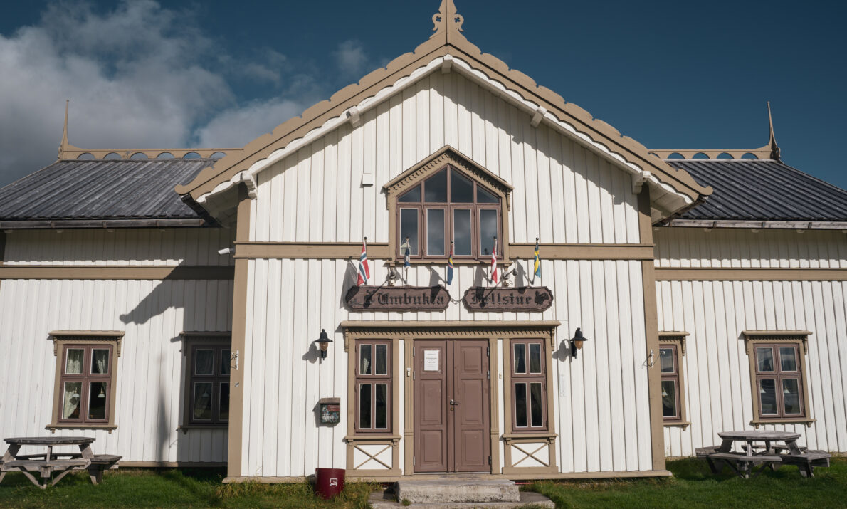 Hvitmalt fjellstue med lys, brune kanter. Blå himmel med noen skyer over.