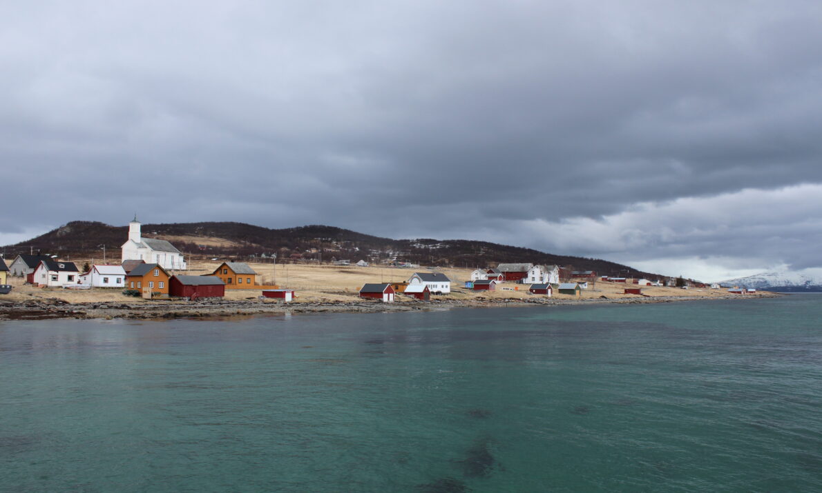 Et kystlandskap med klart, turkist hav i forgrunnen og en rekke spredte bygninger langs strandlinjen. Husene er hovedsakelig lave trebygninger i lyse og røde farger. Landskapet bak består av gress- og lyngkledde åser under en overskyet, grå himmel.