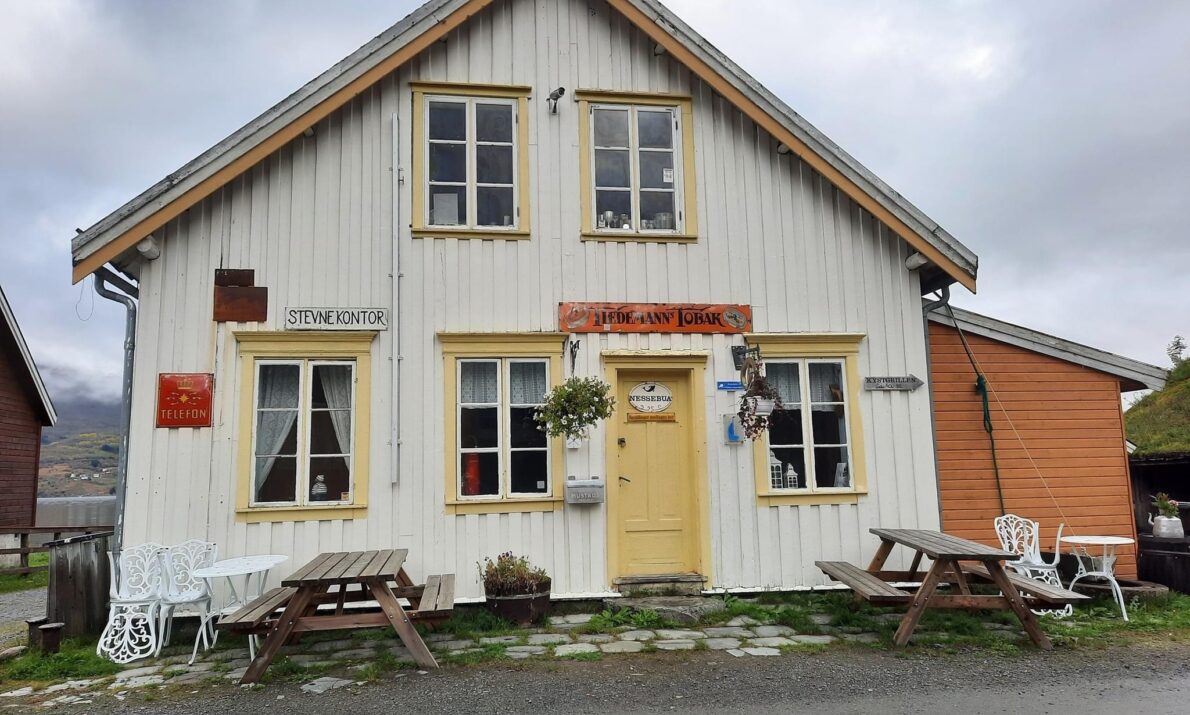 "Et hvitt, toetasjes trehus med gult panel og røde detaljer, omgitt av benker og en grusvei, med fjell i bakgrunnen under en overskyet himmel."