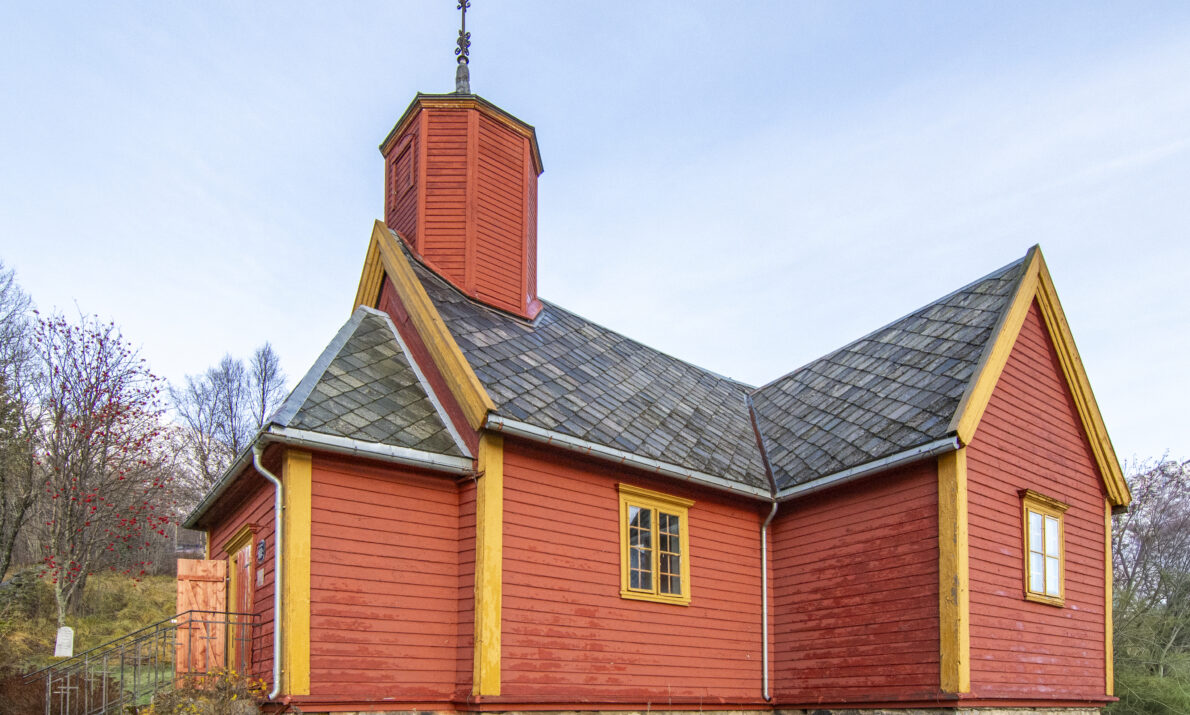 En liten, rød trekirke med gule hjørnelister og steinsokkel, omgitt av gress og trær. Bygget har en lav tårnkonstruksjon med spir og skifertekket tak. Himmelel er lys og klar.