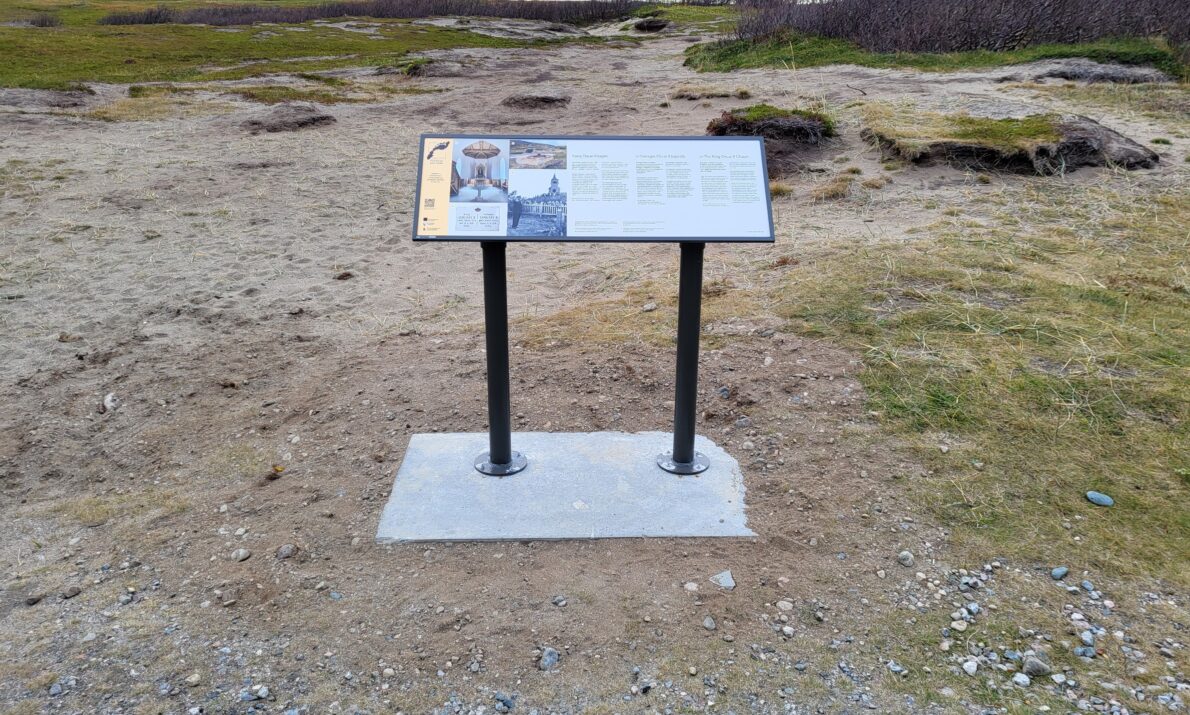 "Et informasjonsskilt på en ståramme i et åpent, karrig landskap med gras og fjell i bakgrunnen."