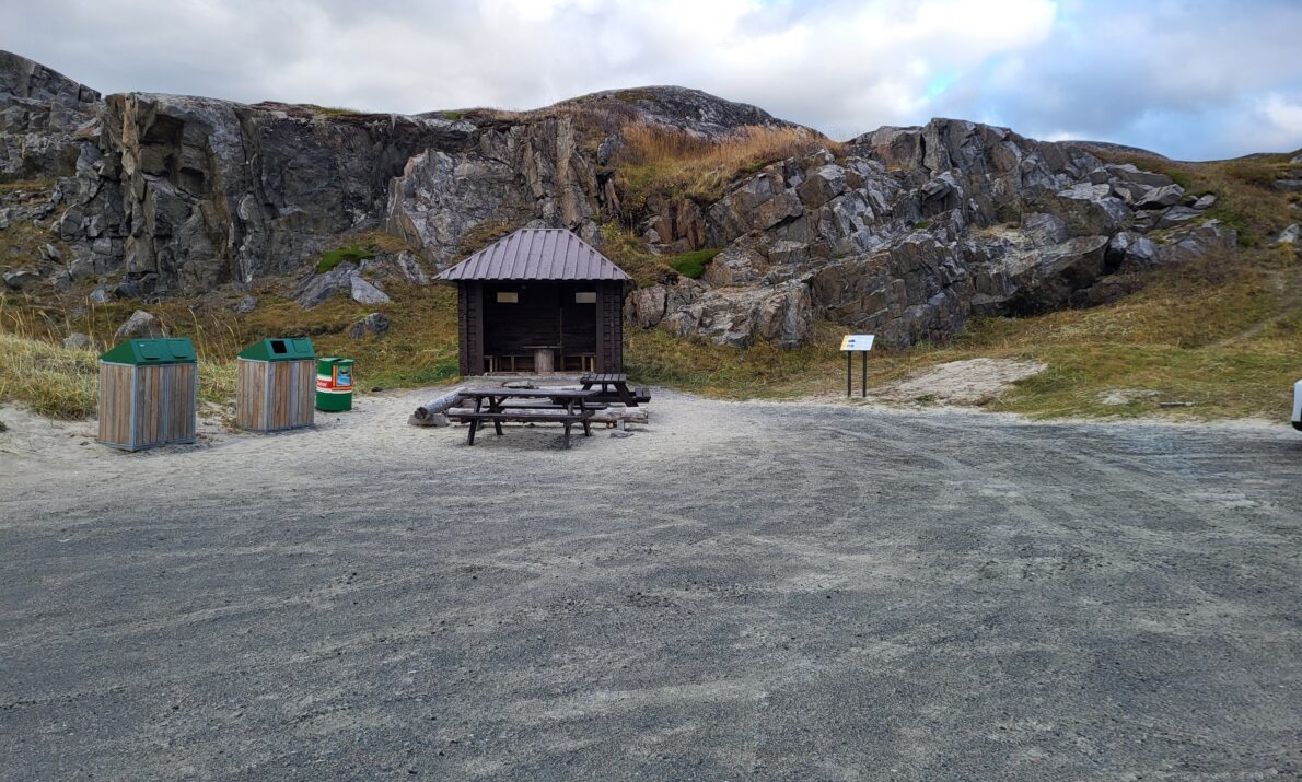 «Et friluftsområde med en åpen, gruslagt plass omgitt av fjell og lav vegetasjon. Midt i bildet står et lite overbygd gapahuk‑lignende bygg med sitteplasser, og foran det står et piknikbord. Til venstre står tre avfallsbeholdere, og til høyre sees en informasjonstavle langs stien. Himmelen er overskyet.»