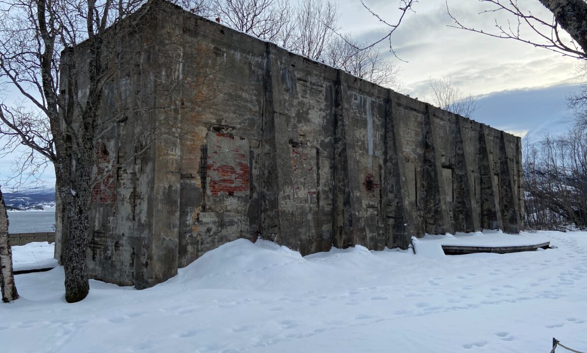 En ruin av et industribygg. Foran noen tre og snø på bakken. Blå himmel med skyer over.