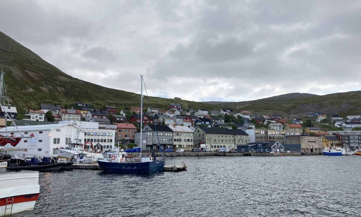 Bilde av Honningsvåg fra sjøen. Sjø i forgrunnen, noen båter og så tett bebyggelse. En ås i bakgrunnen og en himmel med tett skydekke.