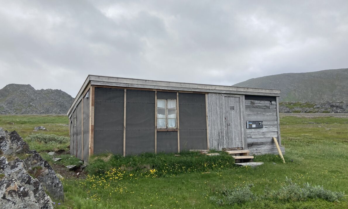 Enkel hytte under rekonstruksjon i midten av bildet. Gress på bakken rundt. Fjell i bakgrunnen og himmel med tett skydekke.