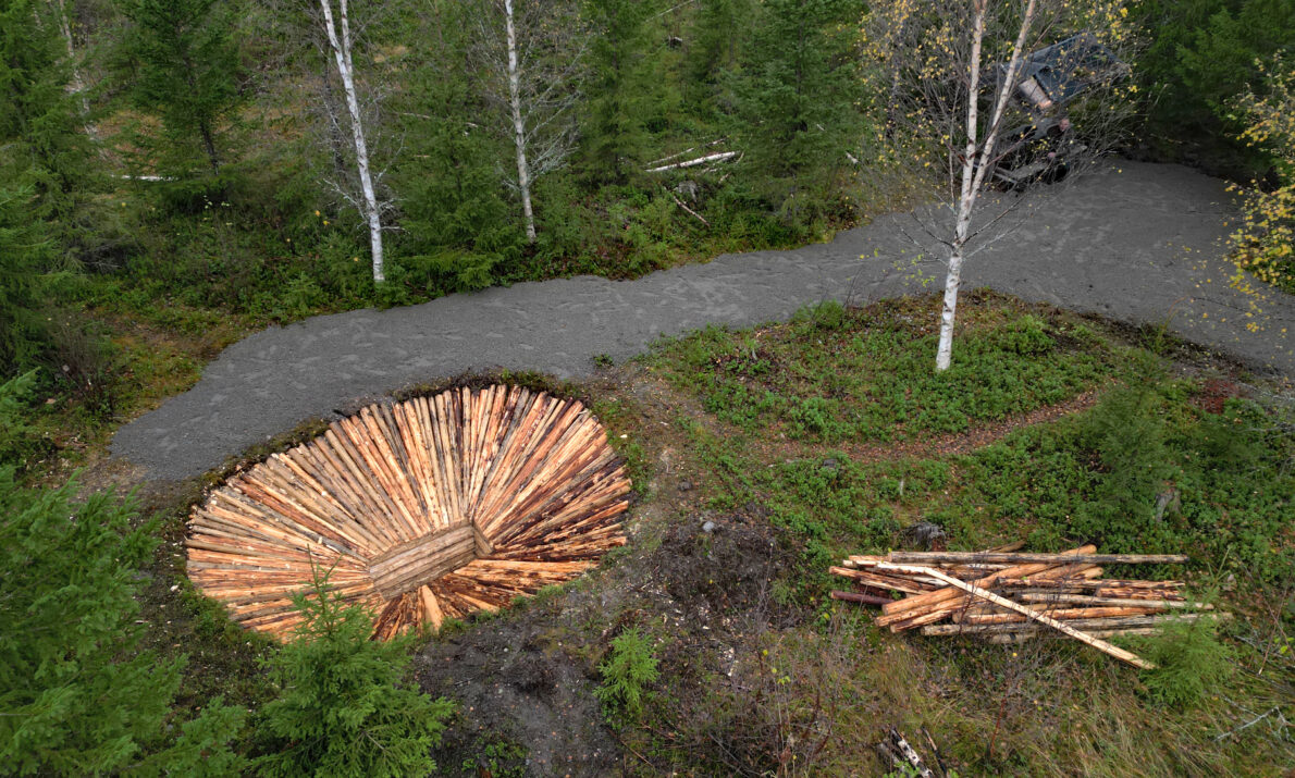 Ser ned på rekonstruert fangstgrop med tømmer. Grusvei bak og noen tømmerstokker til høyre. Vegetasjon og noen trær rundt.