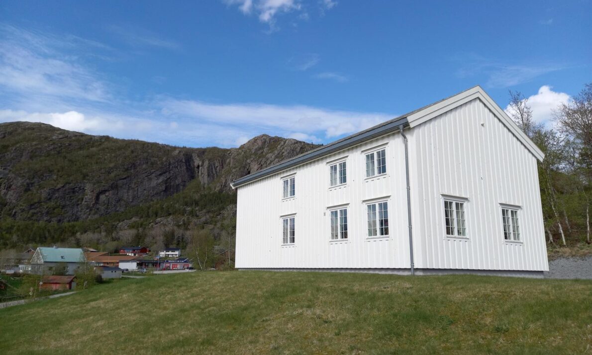 Liten bakketopp med grønt gress. På toppen et hvitt, gammelt hus. Til venstre, litt lengre bort, noen bygninger. Over dem en ås. Blå himmel med noen få hvite skyer.