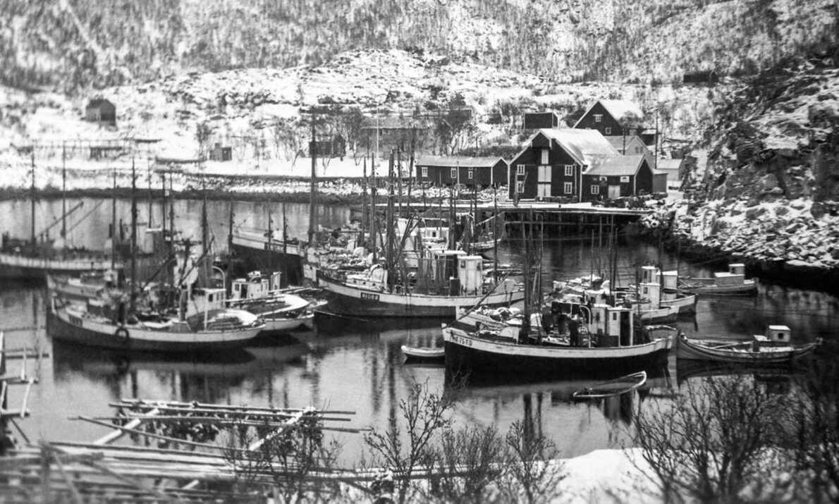 Svart‑hvitt fotografi av en liten havn med flere fiskebåter fortøyd ved brygger. Rundt havnen ligger snødekte bygninger og terreng, omkranset av vinterkledde fjellsider.