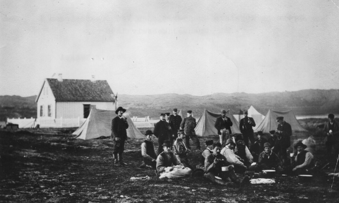 Svart hvitt bilde. Arbeidere står og sitter på en vidde foran noen telt. Hvitt hus i bakgrunnen. Fjell lengst bak.