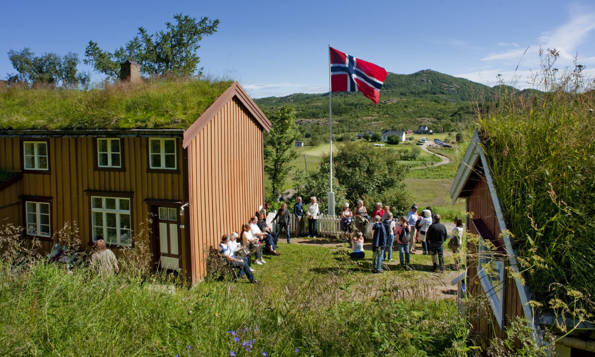 Gammel, okergult hus med torvtak i grønne omgivelser. Et norsk flagg er heist i hagen og en rekke mennesker er samlet utenfor huset. En ås i bakgrunnen og blå himmel.