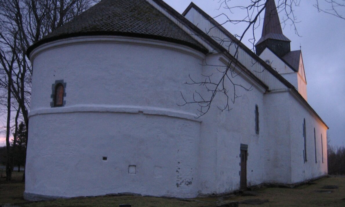En hvit murkirke fotografert i skumringen. Bygningen har en rund apsis, smale vinduer og et høyt tårn med spiss topp. Trær uten løv omkranser området, og kirken står på en gresskledd tomt med dempet, blålig kveldshimmel i bakgrunnen.