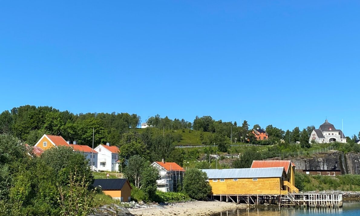 Sommerlig kystlandskap med klart blå himmel. Flere gule og hvite trehus ligger langs vannkanten, med grønne trær og busker rundt. Vannet er stille og speiler bygningene og terrenget. Bakenfor ligger et lite kulturlandskap med spredte hus og gresskledde bakker.