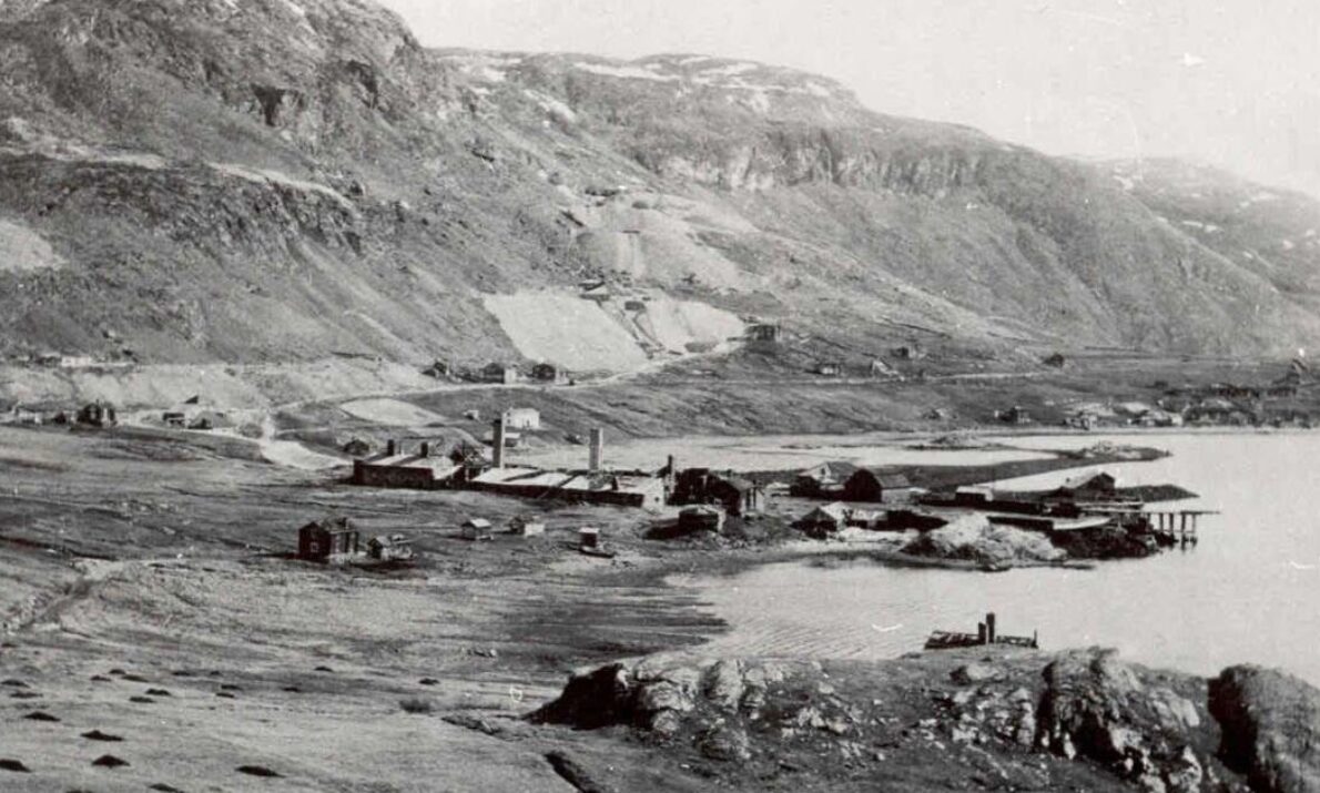 Historisk svart‑hvitt‑foto av et lite industristed ved en fjord, omgitt av fjell. Flere lave bygninger, fabrikkpiper og bryggeanlegg ligger spredt langs vannkanten. Landskapet rundt består av åpne vidder og nakne fjellsider.