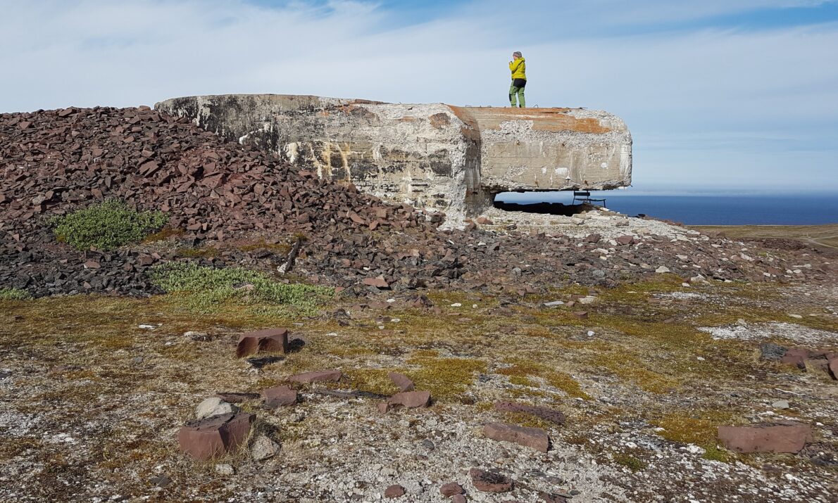 «En åpen, steinete vidde med lav vegetasjon og ujevnt terreng. Midt i bildet ligger en stor, rektangulær betongstruktur som ser ut som en eldre bunkerlignende konstruksjon. En person i en gul jakke står oppe på konstruksjonen og skuer utover området. I bakgrunnen er himmelen lys og delvis skyet, med antydning til hav i horisonten.»