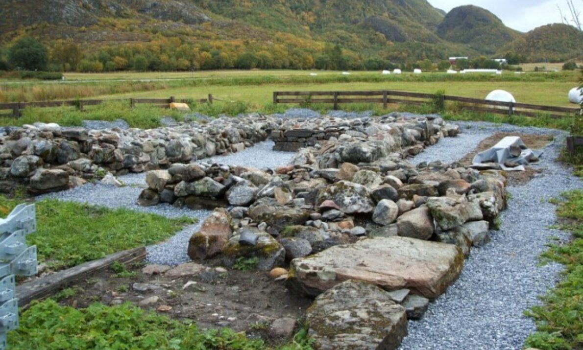 Ruiner av en kirke i forgrunnen. Grønn eng lenger bak og et grønnkledt fjell i bakgrunnen. Himmel med skyer lengst bak.