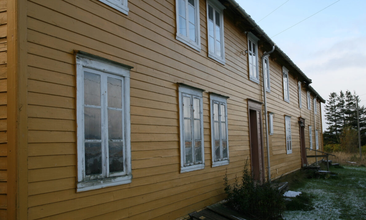 Gammelt, gulmalt hus. Noe gress foran til høyre. Noen trær og blå himmel bak.