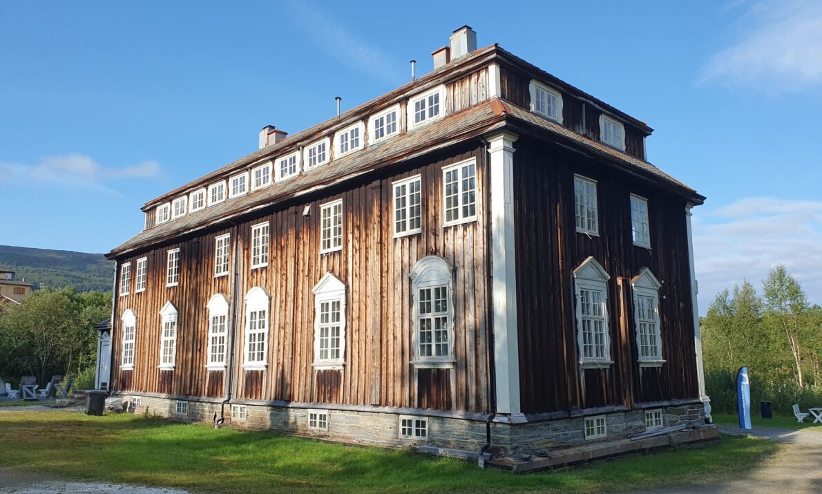 Stort gammel bygg i tre. Malt brunt med hvite karmer. Gress og grus rundt bygget. En ås i bakgrunnen og blå himmel.