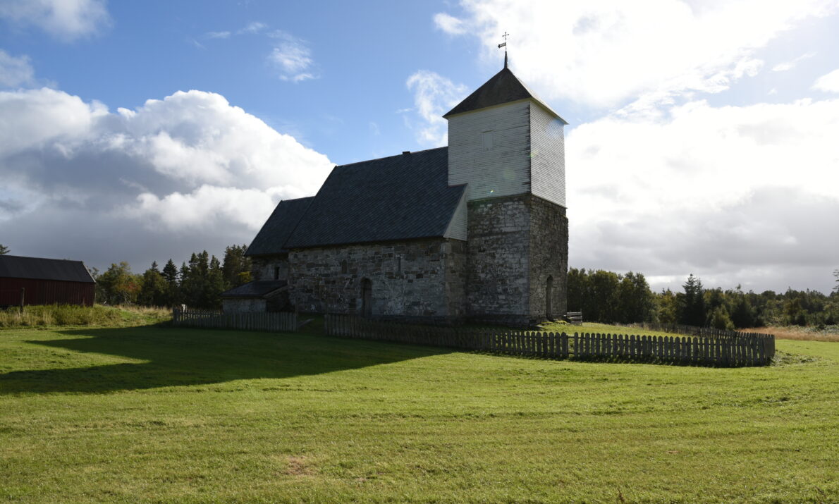 Stor gresslette i forgrunnen og så en gammel, hvit og grå steinkirke i midten. Noen trær bak. Blå himmel med skyer.