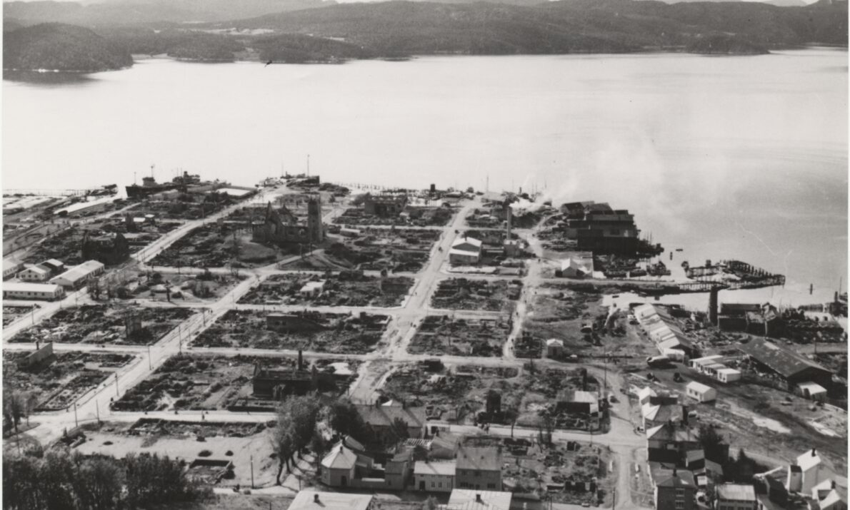 Svart hvitt bilde. Ødelagte bygninger over hele sentrum etter bombing. Vann utenfor og noen åser helt bak.