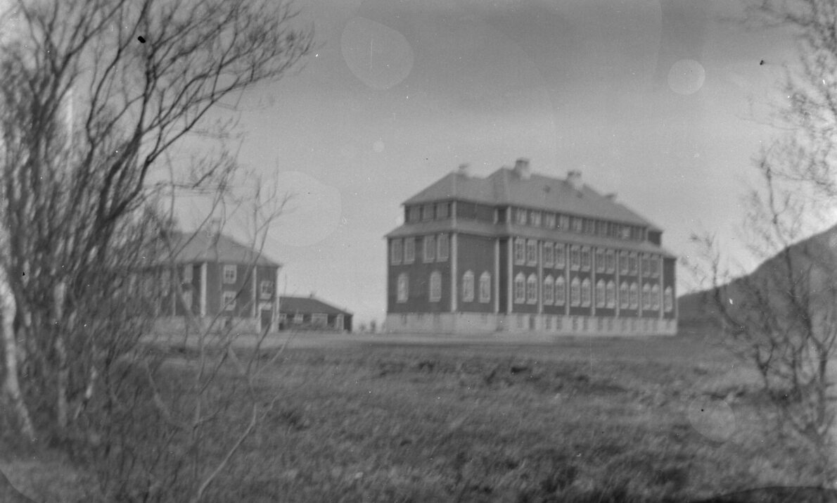 Gammelt svart‑hvitt‑fotografi som viser to store murbygninger i et åpent landskap. Bygningene har flere etasjer, symmetriske vindusrekker og bratte tak. Trær uten løv rammer inn motivet i forgrunnen, og bakken foran bygningene er gresskledd og ujevn.
