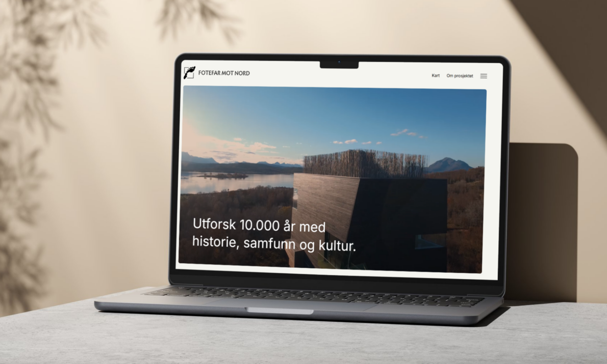 "En bærbar datamaskin som viser nettsiden til Fotefar Mot Nord. På skjermen er det et bilde av et moderne bygningselement med utsikt over et landskap med fjell, vann og skog. Over bildet står teksten: 'Utforsk 10.000 år med historie, samfunn og kultur.' Nettsiden har en navigasjonsmeny øverst med lenker som 'Kart' og 'Om prosjektet'."