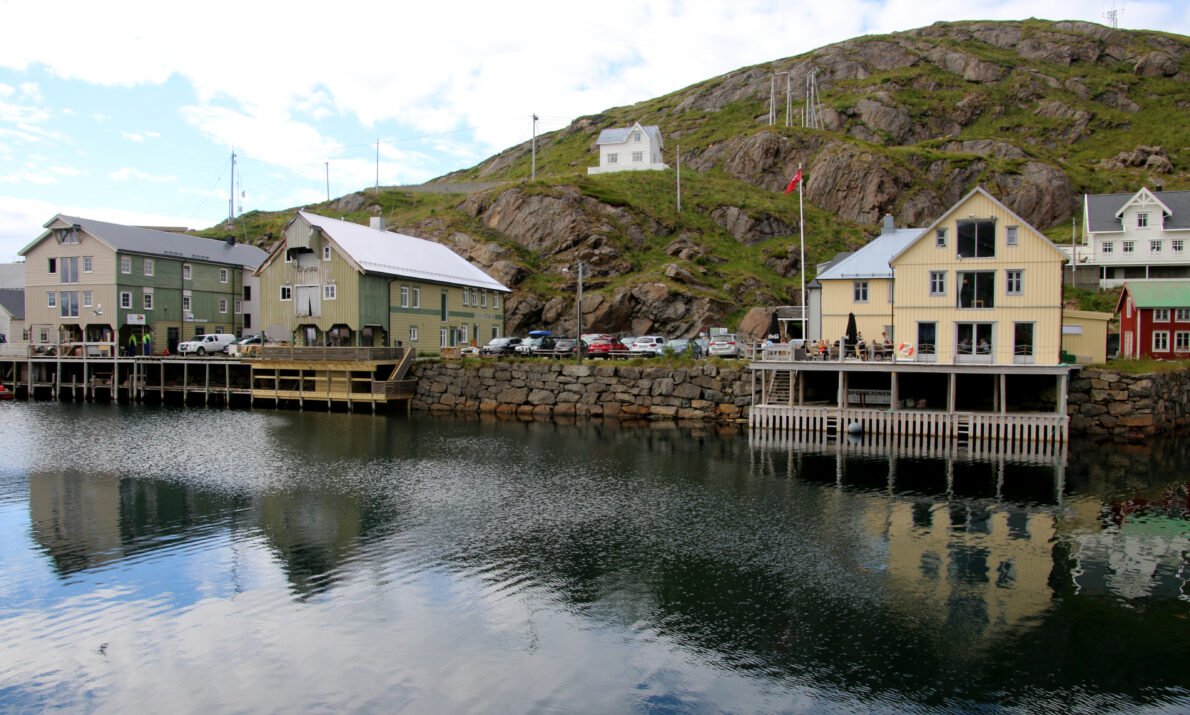 Noen gamle bygninger i et nedlagt fiskevær sett fra sjøen. En lien gresskledt høyde bak et et lite hvitt hus. Blå himmel med skyer over.