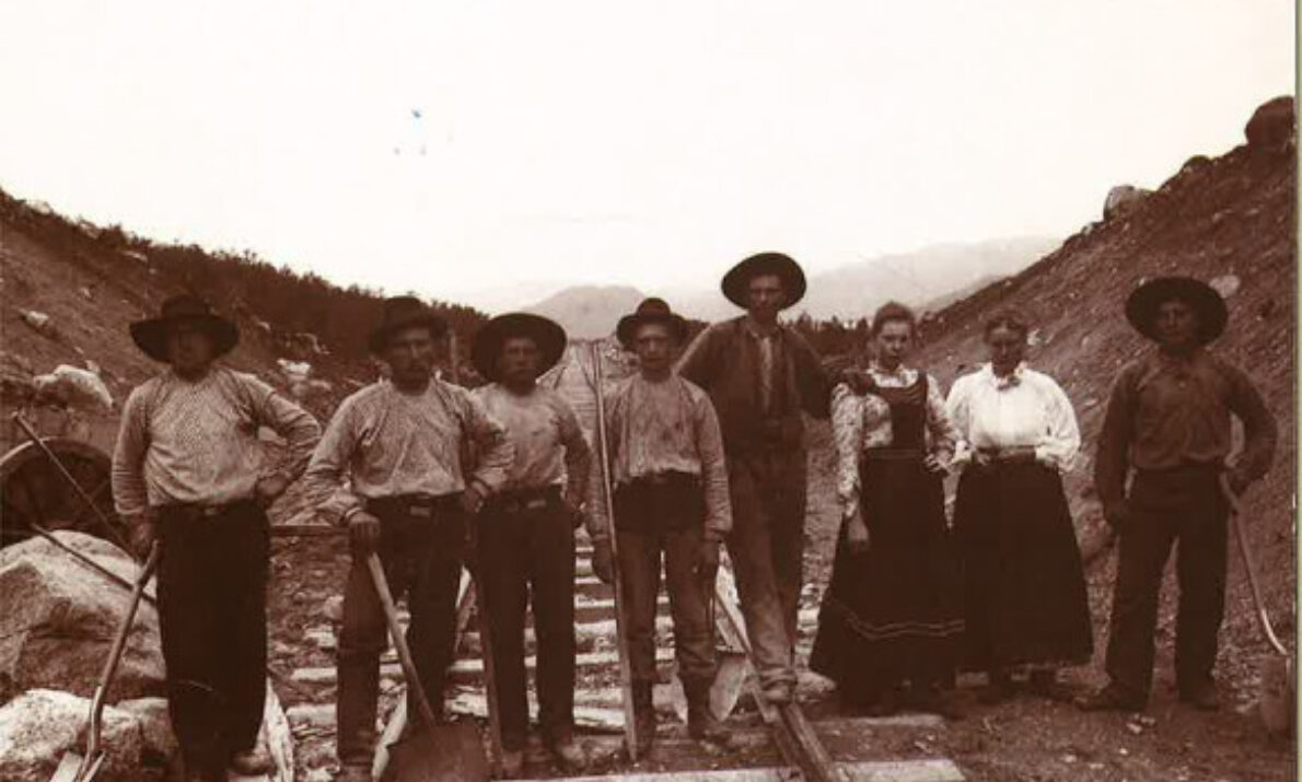 Svart hvitt bilde. Menn og kvinner med hakker og annet utstyr står på jernbaneskinner. Steiner og grus rundt. Fjell i bakgrunnen. Hvit himmel.