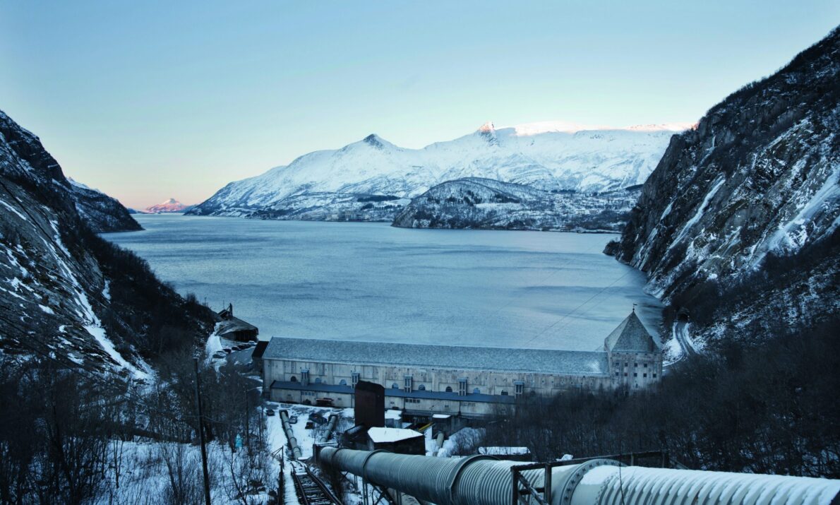 Kraftverk i forgrunnen med et stort rør som går opp mot fotografen. Nederst og innerst i en fjord ligger kraftverket. Utsikt over fjorden og mot snødekte fjell.