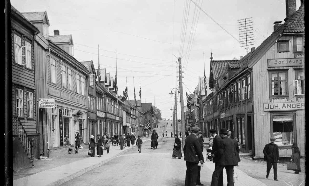 Svart hvitt bilde. Bygate med hus på begge sider. Flere mennesker går i gaten. Hvit himmel over.