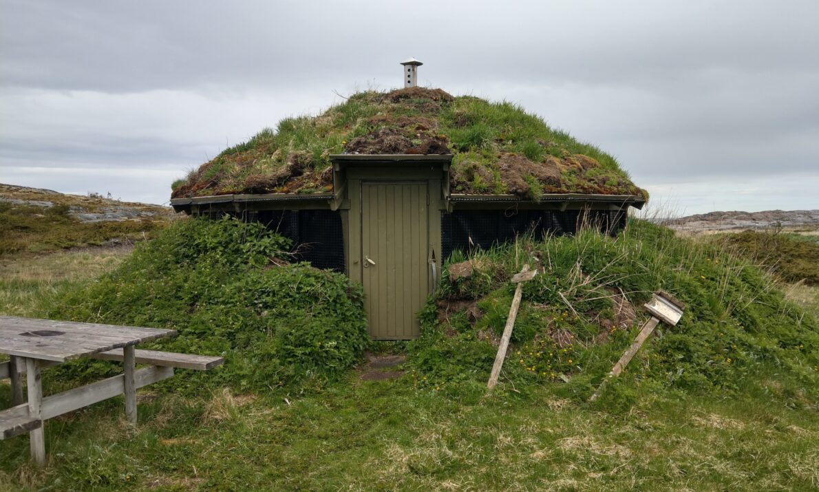 En liten, rund bygning med torvtak og en liten dør. Gress rundt. En benk i tre til høyre og et par skilt som nesten har falt ned. Himmel med tett skydekke over.