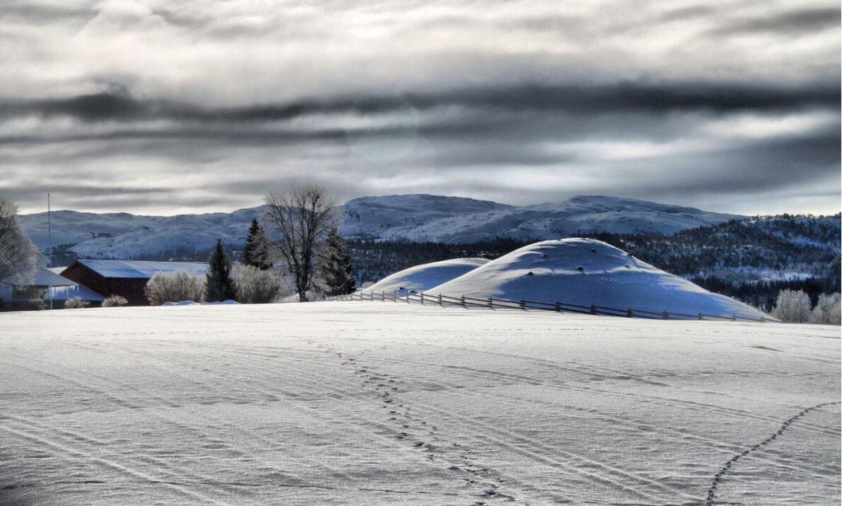 Snødekt slette og to gravhauger i enden, også dekket av snø. Bak der utsikt mot landskap med trær, neon få bygninger, fjell og himmel med skyer.