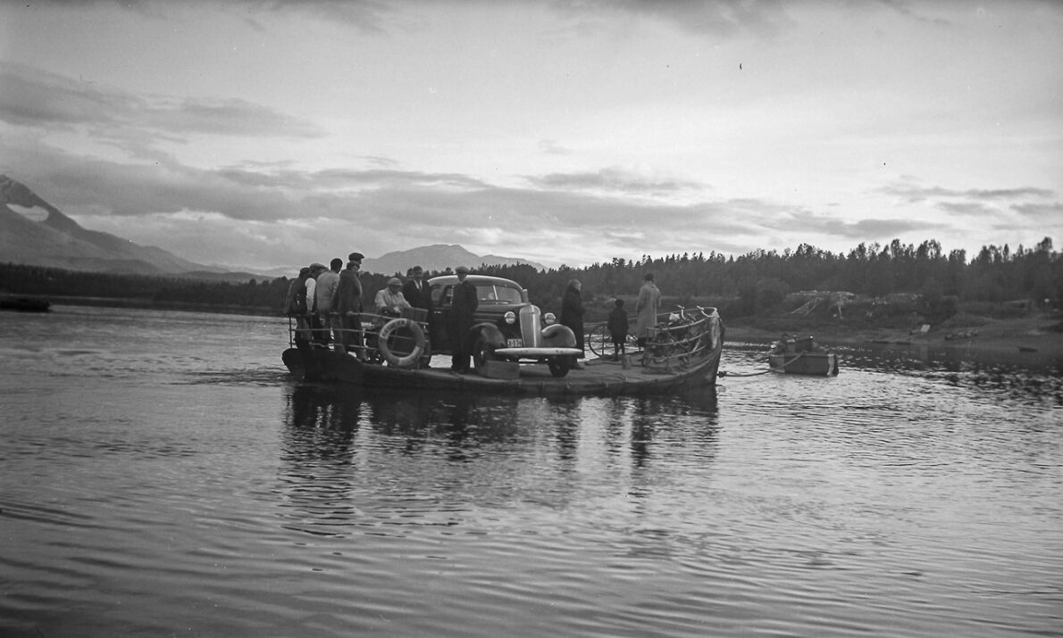 Et svart‑hvitt fotografi som viser en liten ferge eller lekter på et stille vann. Farkosten frakter et kjøretøy og flere personer står tett samlet om bord. I bakgrunnen sees skogkledde åser og fjell under en overskyet himmel.