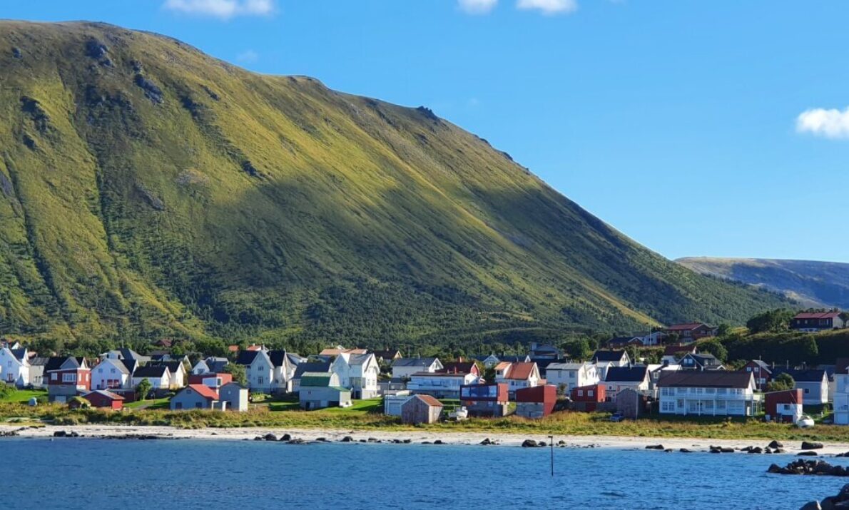 En landsby sett fra havet. Hav foran, en klynge med hus, et fjell kledt i grønt bak og så blå himmel.