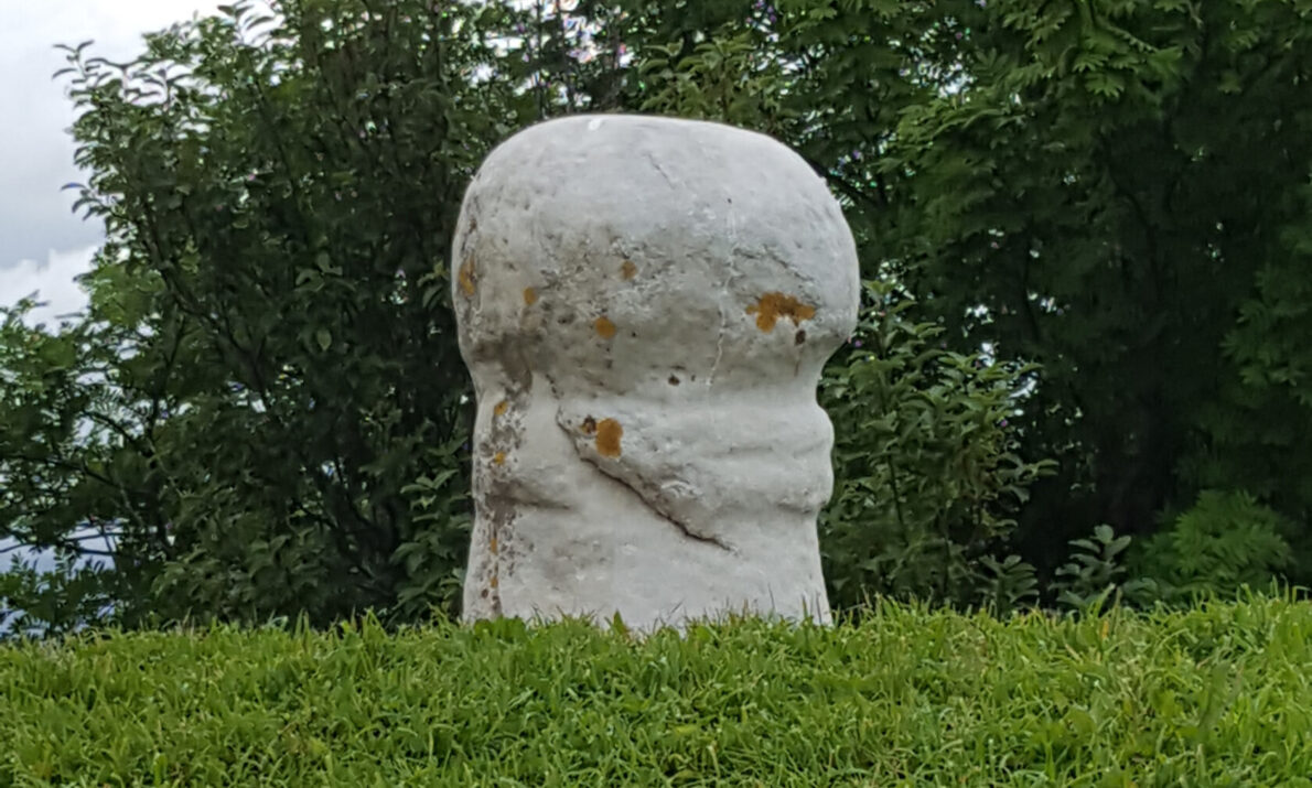 Liten, lys steinskulptur med avrundet form står på en gresskledd bakke, omgitt av trær og under en overskyet himmel.