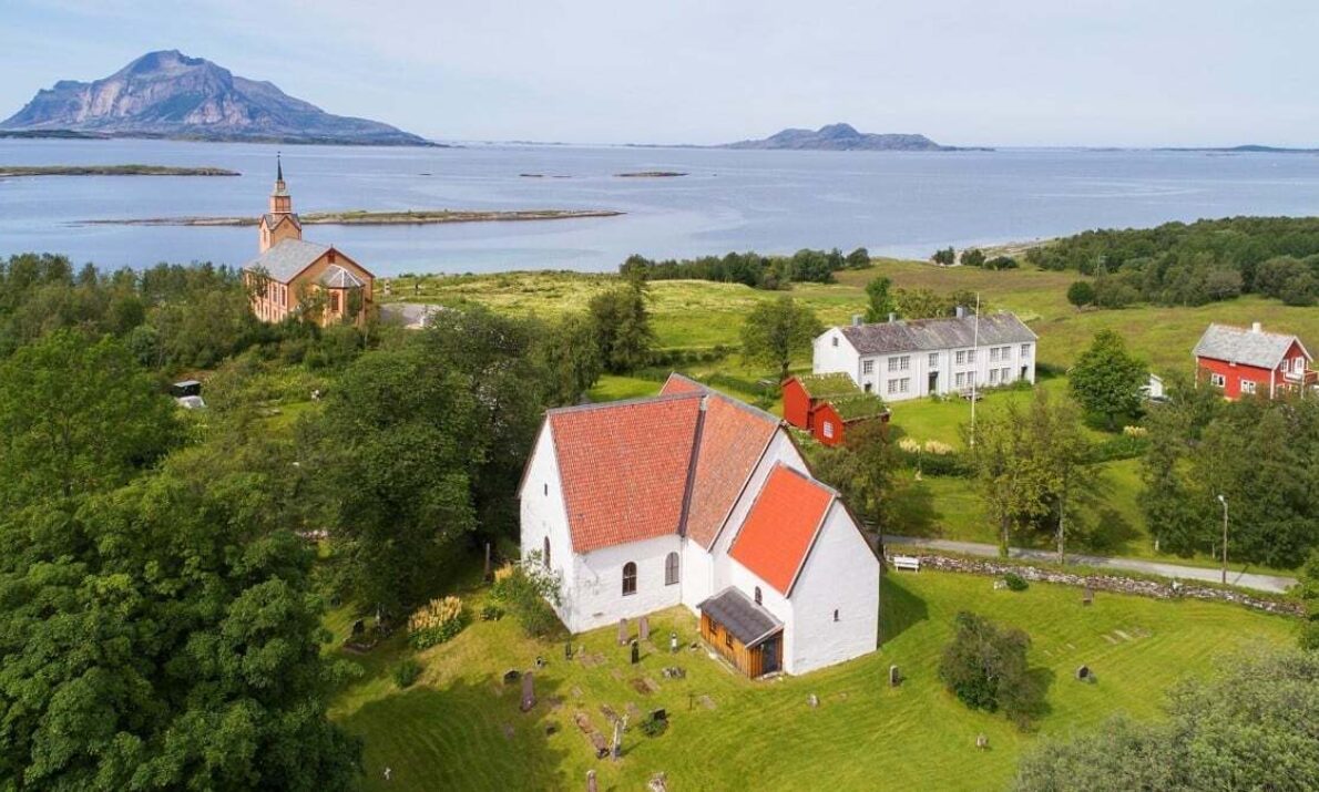 Flyfoto av et grønt kystlandskap med flere bygninger, inkludert en hvit kirke med rødt tak i forgrunnen og en større steinkirke lenger bak. Rundt bygningene ligger åpne jorder, trær og spredte hus. I bakgrunnen skimtes havet og flere fjellformasjoner.