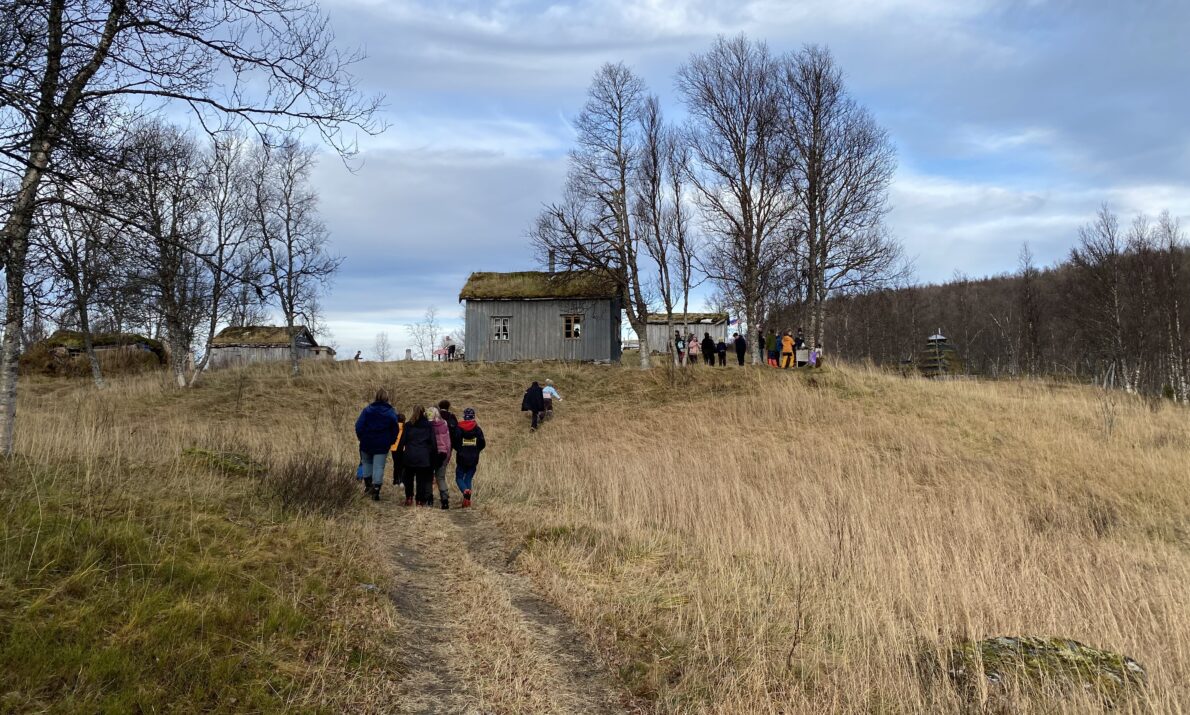 Gamle trehus på en liten høyde med noen trær rundt. Bare på en sti på vei opp mot husene. Lettskyet vær og høstfarger i naturen.