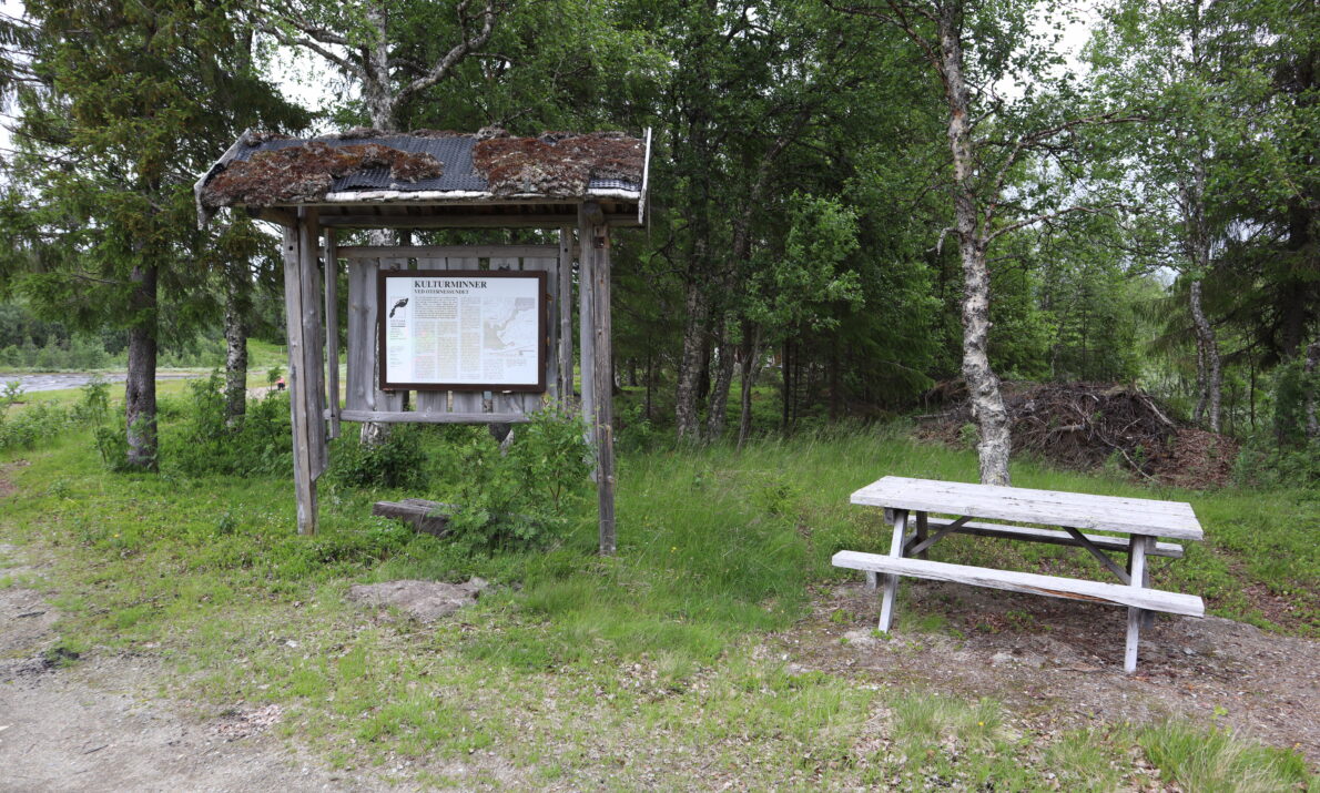 En grusvei forrest og så noe gress. Det står en trebenk til høyre og et stort skilt med treramme og tak til venstre. Skog bak.