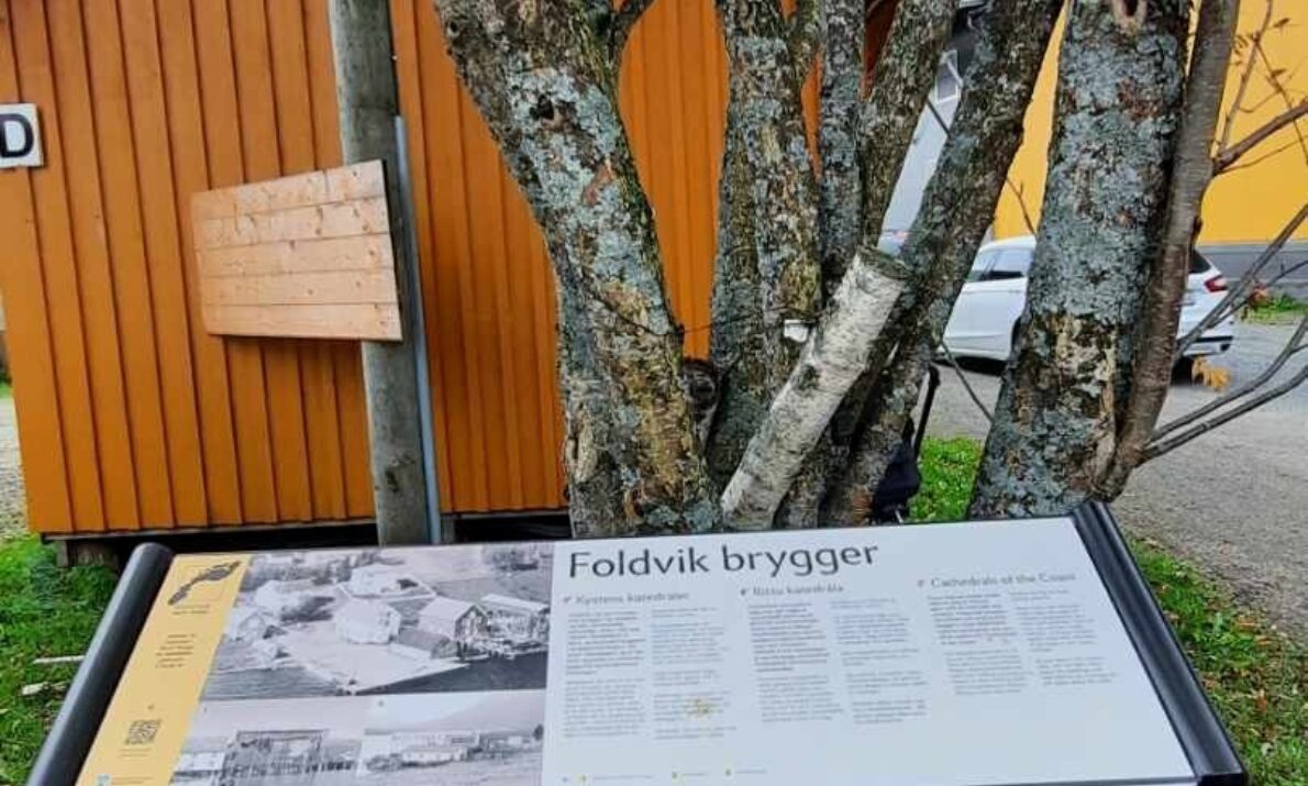 Stort informasjonskilt foran tre og en orangemalt trebrygge