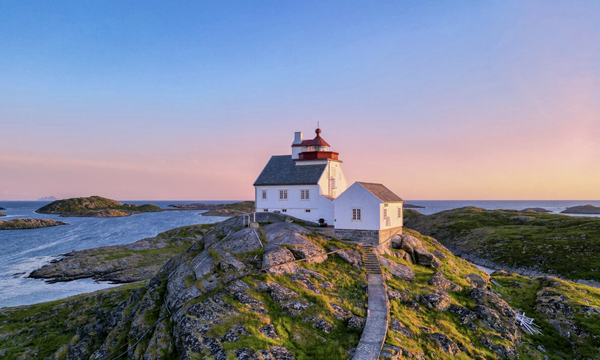Et hvitt fyrhus med rødt tak står på en liten, steinete øy omgitt av hav. En smal tresti leder opp til bygningen. Landskapet badet i varmt lys fra solnedgangen, og små øyer ligger spredt i bakgrunnen.