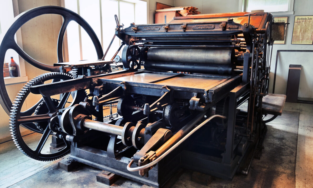 En stor, gammel trykkpresse i et museumsmiljø. Maskinen er laget av mørkt metall med store tannhjul, valser og et svinghjul på venstre side. Den står i et lyst rom med vinduer bak og trykkerirelaterte gjenstander på veggene.